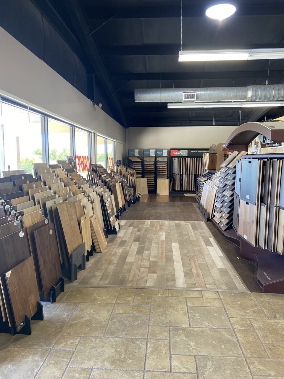 Wide showroom aisle of engineered hardwood flooring samples with a Bella Cera premium hardwood display sign